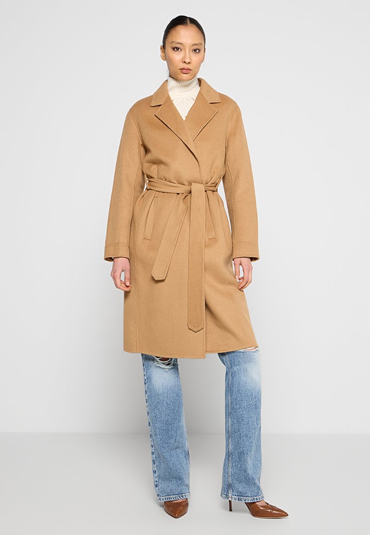 Kobieta w beżowym płaszczu z paskiem nałożonym na białą golfową bluzkę i podartych niebieskich dżinsach, stojąca na tle gładkiej białej ściany.