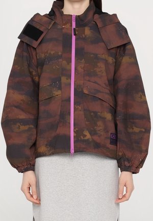 Osoba ubrana w brązową i ciemnopomarańczową kurtkę moro z jasnoróżowym, frontowym zamkiem błyskawicznym i elastycznymi mankietami, stojąca na jednolitym tle.