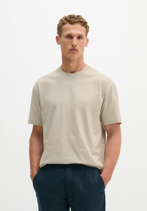 Jonge man met kort krullend haar, die een beige kort mouwen overhemd en donkerblauwe broek draagt, met handen in de zakken tegen een effen achtergrond.