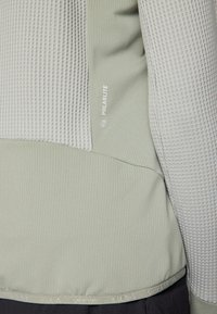 Top atletico grigio chiaro con texture, caratterizzato da un motivo a righe e sezioni lisce; presenta il marchio "POLARLITE" in lettere bianche sottili.