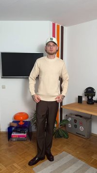 Beżowy sweter dzianinowy z prążkowanymi mankietami, zestawiony z brązowymi spodniami i błyszczącymi ciemnymi butami. Dodatki to biała czapka i tatuaże na ramionach.