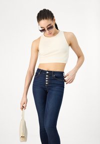 Κρέμ ribbed κοντό τοπ με ψηλό λαιμό και σκούρους μπλε ψηλόμεσους τζιν με πέντε μεγάλους ασημένιους κόμπους. Αξεσουάρ περιλαμβάνουν γυαλιά ηλίου.