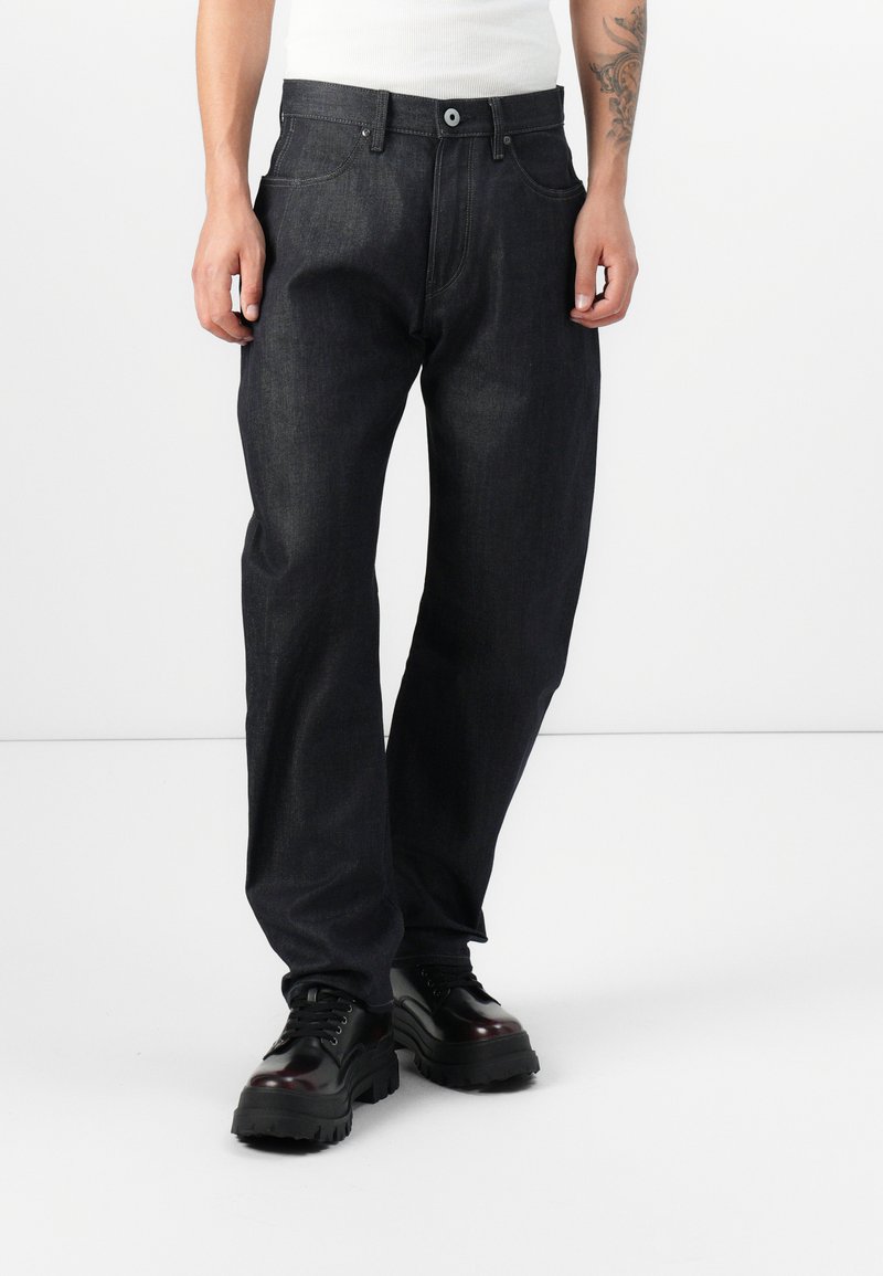 Dark denim jeans with a straight fit, front pockets, and a button fly closure. Worn with black chunky-soled shoes. Smooth texture.