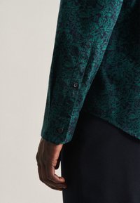 Camicia verde con motivo floreale. Presenta un polsino con bottoni e un tessuto testurizzato. Si adatta comodamente grazie a un taglio sartoriale.