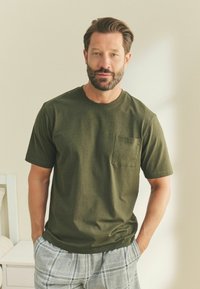 Olivegrünes Baumwoll-T-Shirt mit kurzen Ärmeln und einer Brusttasche, kombiniert mit hellgrauen karierten Shorts. Einfaches, lässiges Design.