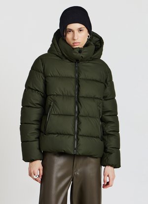 Person in einer dunkelgrünen Steppjacke mit hohem Kragen und schwarzer Mütze, kombiniert mit braunen Lederhosen vor einem einfarbigen Hintergrund.