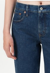 Jean en denim bleu foncé avec taille haute, conception à cinq poches, rivets en cuivre et détails de couture visibles. Texture lisse et matériau résistant.