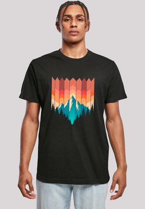 Young man wearing black T-shirt with colorful graphic of a mountain and pine trees against a sunset sky, light wash jeans.