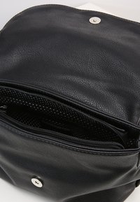 Bolso de cuero negro con una textura suave, cierre de cremallera y forro de lunares. Presenta un botón de presión plateado en la parte frontal.