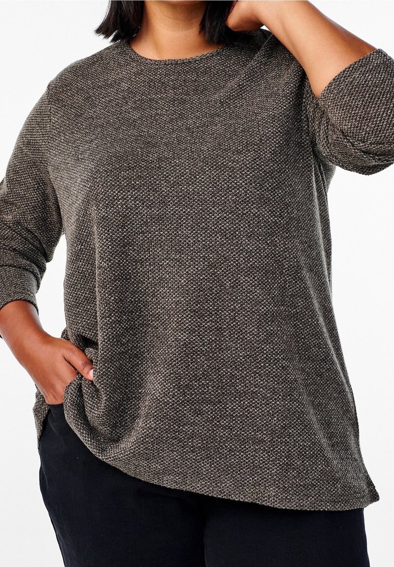 Femme à la peau mate portant un pull gris texturé à manches longues et un pantalon noir, une main dans la poche, l'autre touchant son cou