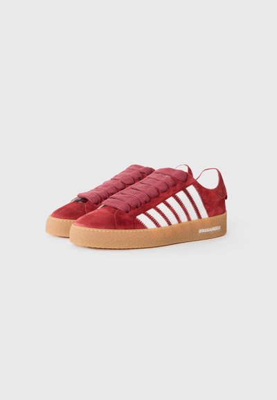 Red suede sneakers with a round toe, featuring thick maroon laces and three white stripes on the side. Rubber gum sole with branding.