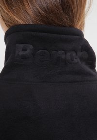 Mustalainen fleece-takki, jossa on korkea kaulus. Takana on kohokuvioitu "Bench." -logo tekstuurin yksityiskohtana. Liukas, pehmeä kangas.