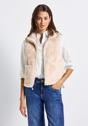 Gilet en fausse fourrure beige avec fermeture éclair, deux poches et col haut, porté sur une chemise blanche à boutons et un jean bleu.