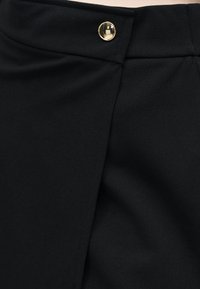 Μαύρη φούστα ψηλόμεση με εμπρόσθιο τύλιγμα και ένα μόνο, λαμπερό χρυσό κουμπί στο ζωνάρι.