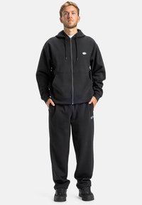 Czarna bluza z kapturem na zamek, z sznurkami, z małym logo. Połączona z pasującymi spodniami dresowymi i czarnymi sneakersami. Miękki materiał, luźny krój.