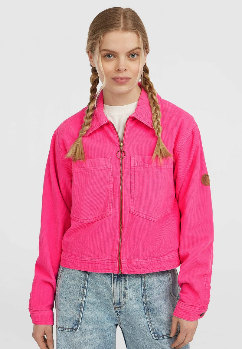 Vrouw met blond haar in twee vlechten, gekleed in een felroze zip-up jack en lichtblauwe high-waisted jeans tegen een effen achtergrond.