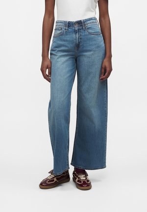 Personne portant un jean large bleu clair, un haut blanc et des baskets bordeaux avec des lacets beige, debout devant un fond blanc uni.