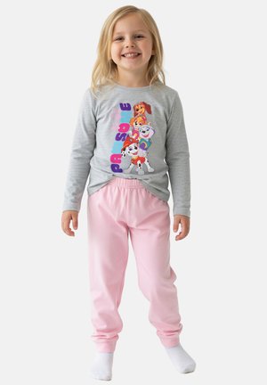 Giovane ragazza sorridente che indossa una maglietta grigia a maniche lunghe Paw Patrol e pantaloni rosa larghi, in piedi davanti a uno sfondo bianco.