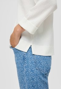 Valkoinen tekstuurilla varustettu pusero, jossa on kolmen neljänneksen hihansuut, yhdistettynä vaaleansinisiin kuviollisiin farkkuihin, joissa on pyörteisiä kuvioita.