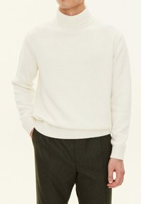 Cremefarbener Strickrollkragenpullover mit gerippten Bündchen und Saum. Kombiniert mit dunkelgrauen Hosen, die einen strukturierten Stoff ohne Muster zeigen.
