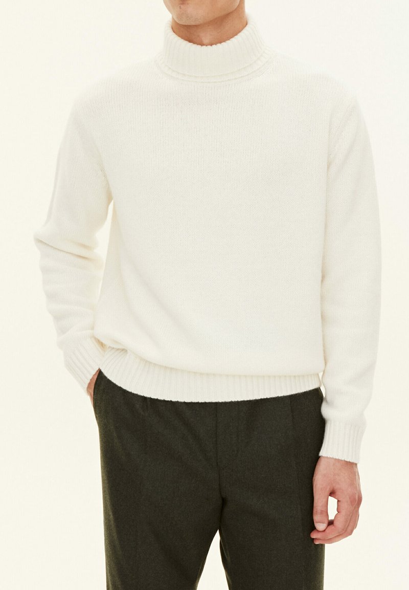 Cremefarbener Strickrollkragenpullover mit gerippten Bündchen und Saum. Kombiniert mit dunkelgrauen Hosen, die einen strukturierten Stoff ohne Muster zeigen.