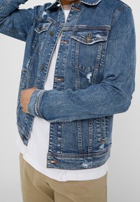 Person wearing a distressed blue denim jacket over a white shirt and beige pants, adjusting the jacket collar with one hand.