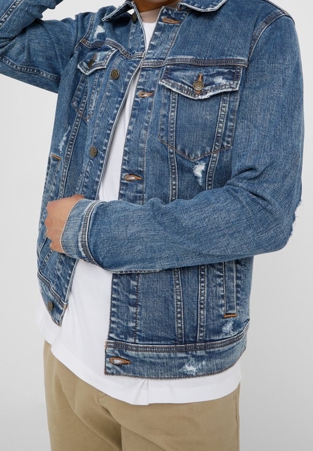 Person wearing a distressed blue denim jacket over a white shirt and beige pants, adjusting the jacket collar with one hand.