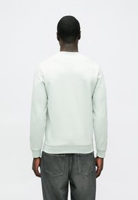 Világosszürke, puha anyagból készült pulóver hosszú ujjakkal és bordázott mandzsettákkal. Sötétszürke farmernadrággal viselve.