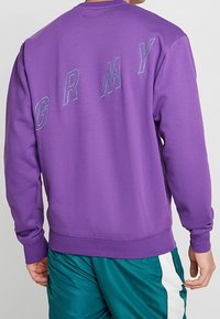 Persona que lleva un sudadera morada con "GRMY" estampado en diagonal en la parte de atrás, combinada con unos pantalones cortos deportivos en color verde azulado con una franja blanca.