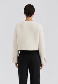 Krótkie białe wełniane kurtki z rozciętymi rękawami i czarnymi wykończeniami, w zestawieniu z czarnymi spodniami o dopasowanym kroju, przedstawione od tyłu. Gładka tekstura.