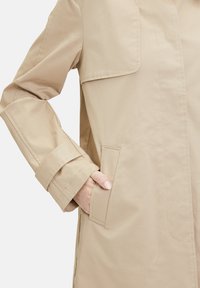 Beige Langarmmantel aus glattem Stoff, mit einer Klapptasche und umgeschlagenen Ärmel. Die Hand ist in die Seitentasche gesteckt.