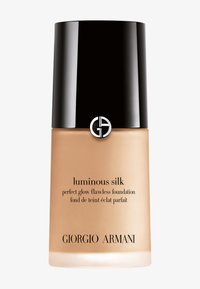 Flacon de fond de teint beige avec un capuchon noir brillant. Verre transparent mettant en valeur le produit. Texte noir comprenant "soie lumineuse" et le nom de la marque.