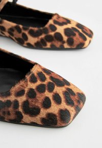 Ballerines en imprimé léopard avec un design à bout carré, fabriquées à partir d'un matériau doux et texturé, avec des taches noires et marron sur une base marron clair.