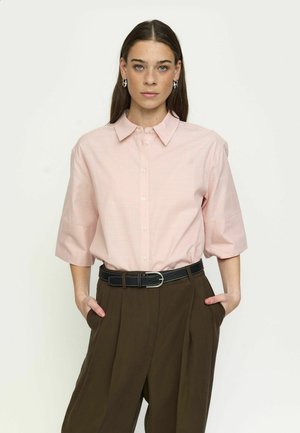 Hosszú sötét hajú nő világos rózsaszín, gombos blúzt és barna, magas derekú nadrágot visel fekete övvel, kezét zsebében tartva.