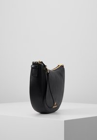 Bolso de cuero negro de forma curva, con herrajes dorados, una cremallera lateral y un asa corta. Superficie texturada con un logo visible.