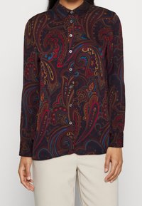 Långärmad skjorta med knäppning i mörk burgundy med ett intrikat paisley-mönster i rött, blått och guld. Har en krage och detaljer med knäppningar på ärmsluten.