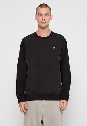 Uomo che indossa una felpa nera a maniche lunghe con scollo rotondo e piccolo logo giallo a forma di uccello sul petto, abbinata a pantaloni beige chiaro, di fronte.