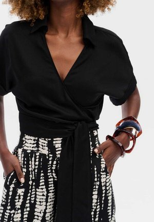 Femme portant un chemisier noir enveloppant et une jupe noire et blanche à motifs, avec des bracelets écaille de tortue et bleus au poignet droit, mains dans les poches.