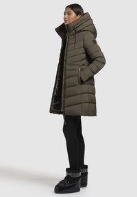 Long olive green puffer coat with a hood, quilted design, zippered front, and black accents. Worn over black leggings and shiny black boots.