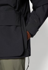 Black waterproof jacket featuring a front pocket, an adjustable hem, and branded detailing on the sleeve, made from a smooth, durable fabric.