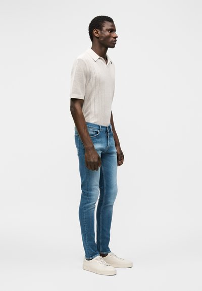 Άνδρας που φοράει μπεζ πλεκτό πόλο με κοντά μανίκια, στενά μπλε τζιν και λευκά αθλητικά, στέκεται προφίλ μπροστά σε απλό φωτεινό φόντο.