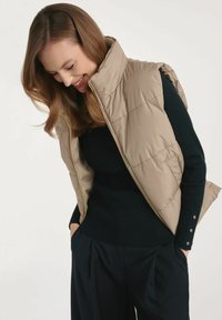 Kobieta z włosami do ramion patrzy w dół, ubrana w beżową kamizelkę puchową na czarną bluzkę z długim rękawem z zapięciem na guziki oraz czarne spodnie.