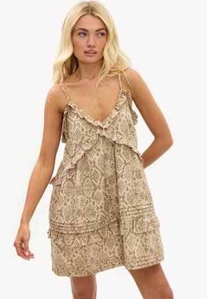 Femme blonde portant une robe beige sans manches à volants avec un motif serpent, regardant sur le côté sur un fond uni.