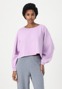 Hanorac scurt de culoare liliac, cu mâneci lungi, decolteu rotund și croială lejeră, asortat cu pantaloni de trening gri, cu talie elastică.