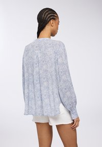 Blusa azzurro chiaro a fantasia con maniche lunghe, orlo arrotondato e dettagli con bottoni. Il tessuto sembra morbido e leggero.