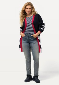 Cárdigan de punto en azul marino con rayas rojas y blancas, con un cierre de botones, llevado sobre una camiseta de rayas azul y blanca y unos pantalones grises con dobladillos deshilachados.