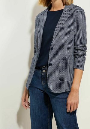 Frau trägt ein marineblau gemustertes Blazer über einem schwarzen Oberteil und blauen Jeans, steht vor einem schlichten hellen Hintergrund.