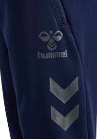Marineblaue Sweatpants mit glatter Textur, versehen mit einem reflektierenden Logo und Chevron-Akzenten in Hellgrau. Einschließlich eines Reißverschluss-Taschen-Details.