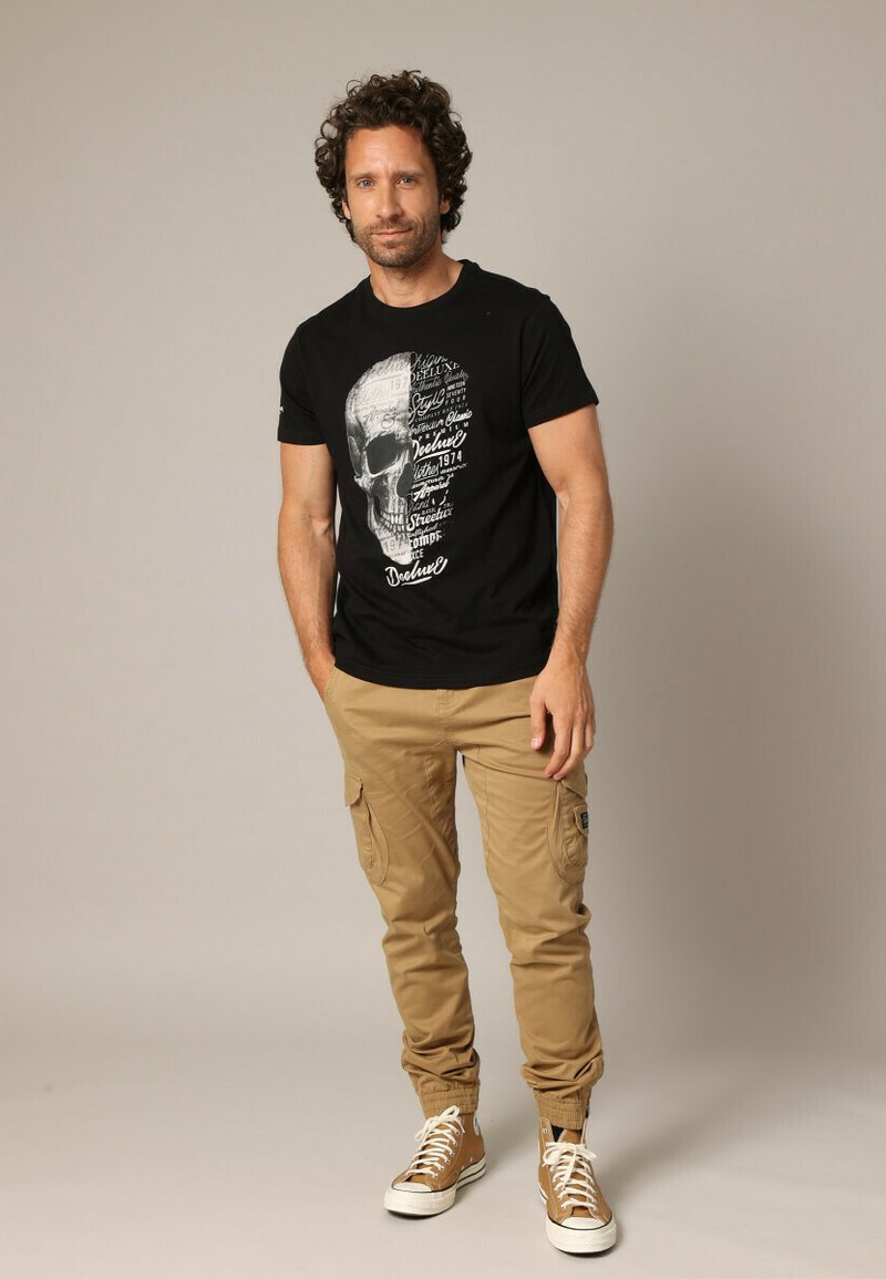 T-shirt noir avec motif de tête de mort et texte, associé à un pantalon cargo beige et des baskets marron. Style décontracté et ajusté.