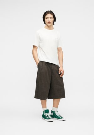 Ung mann med mørkt hår som har på seg en hvit kortermet t-skjorte, løse mørkebrune knebukser, hvite sokker og grønne høy-toppsneakers, stående.